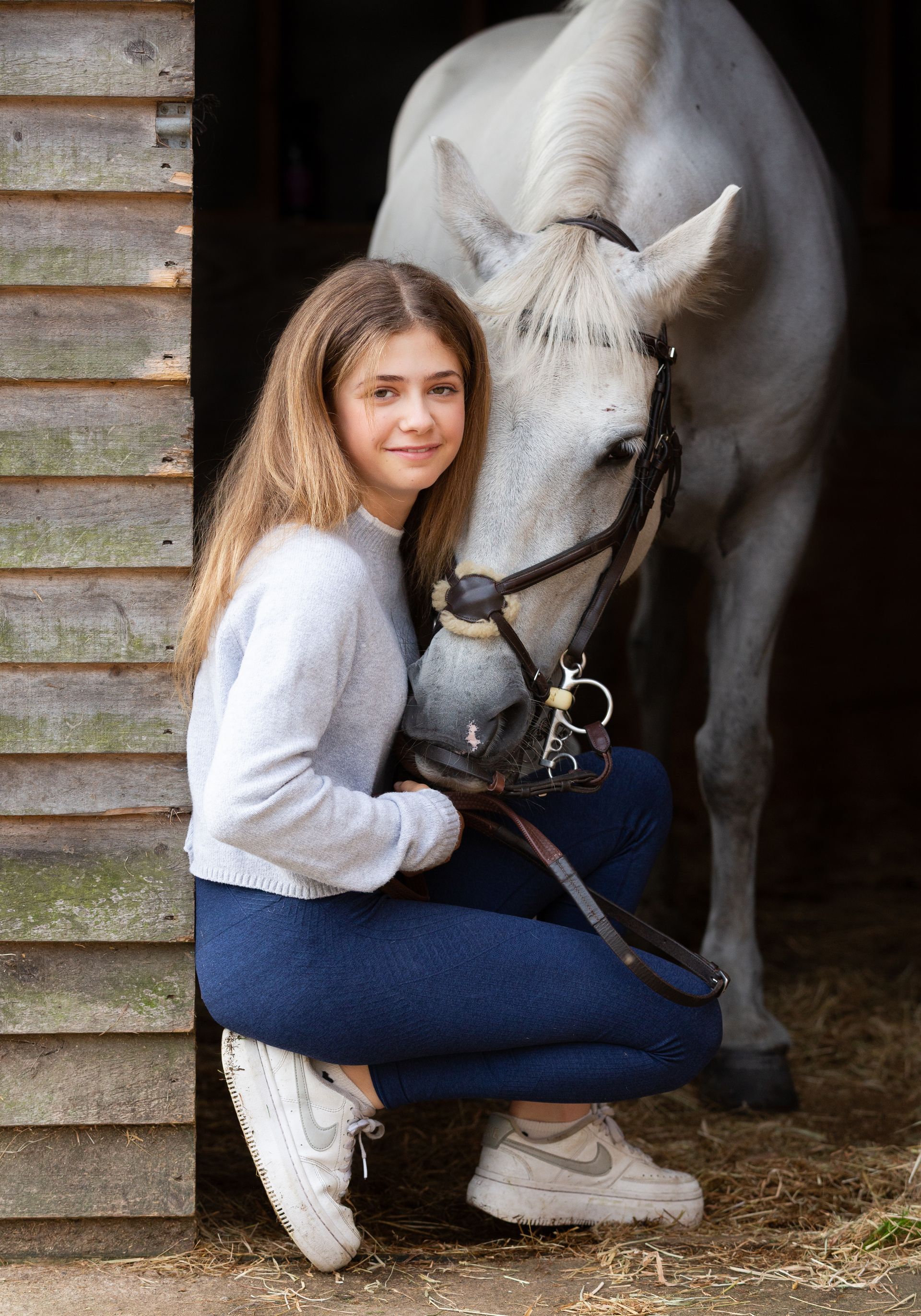 Girl in jeans and sweater kneels next to a white horse in a stable.