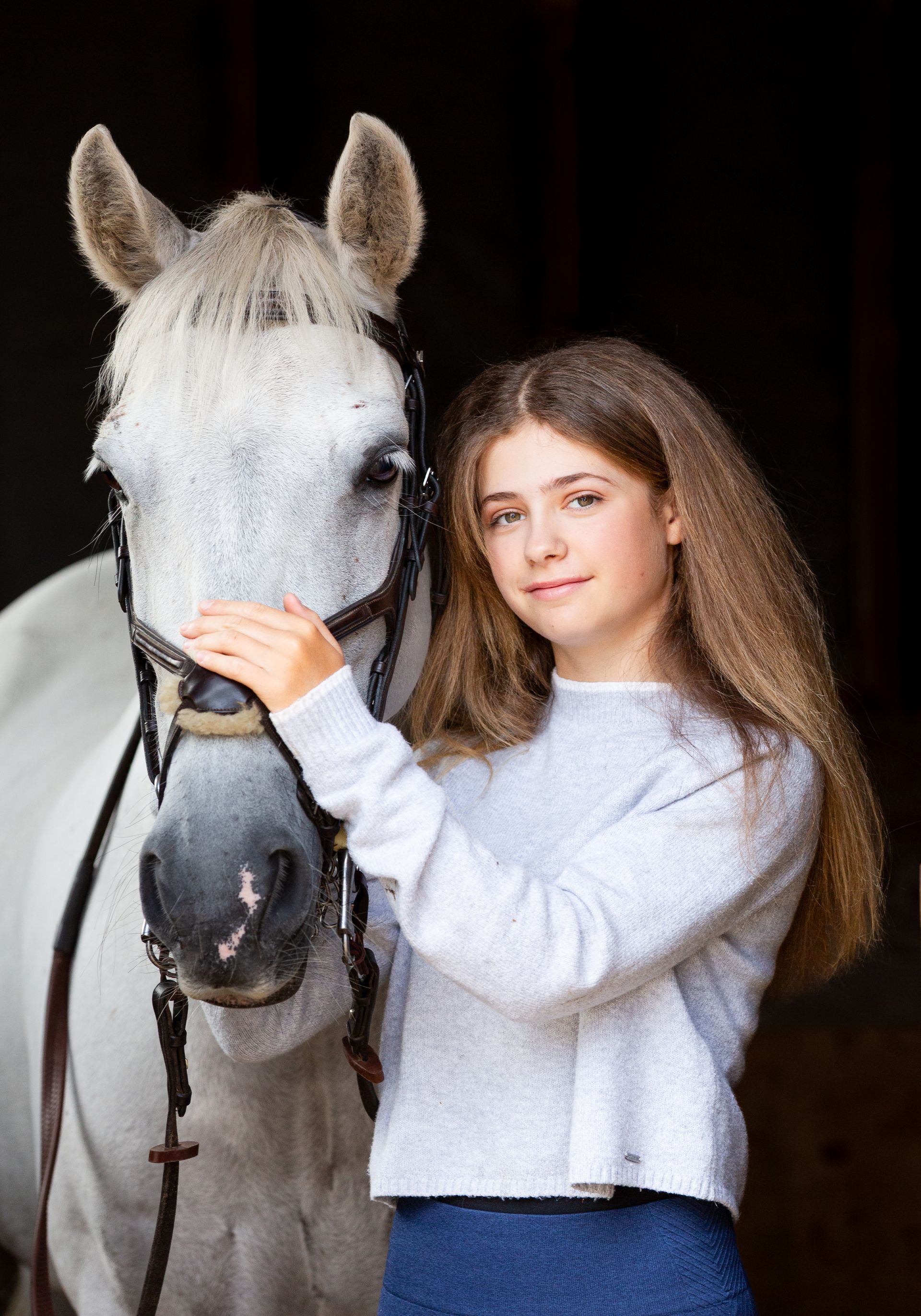 Girl petting a white horse. They're indoors. The girl is wearing a light sweater and smiling.