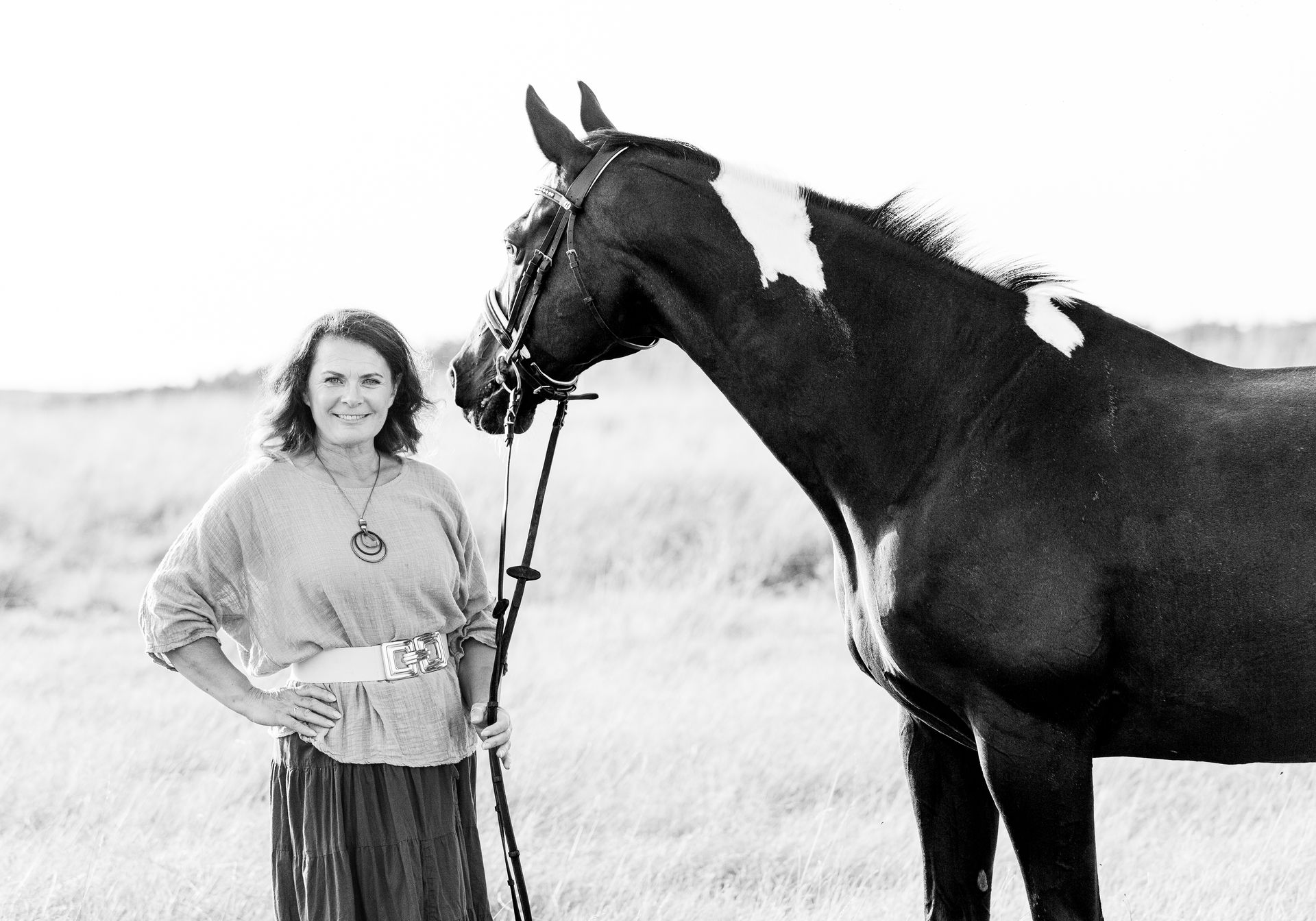 Woman standing with horse in a field. Horse is black and white, woman has hand on hip, both smiling.