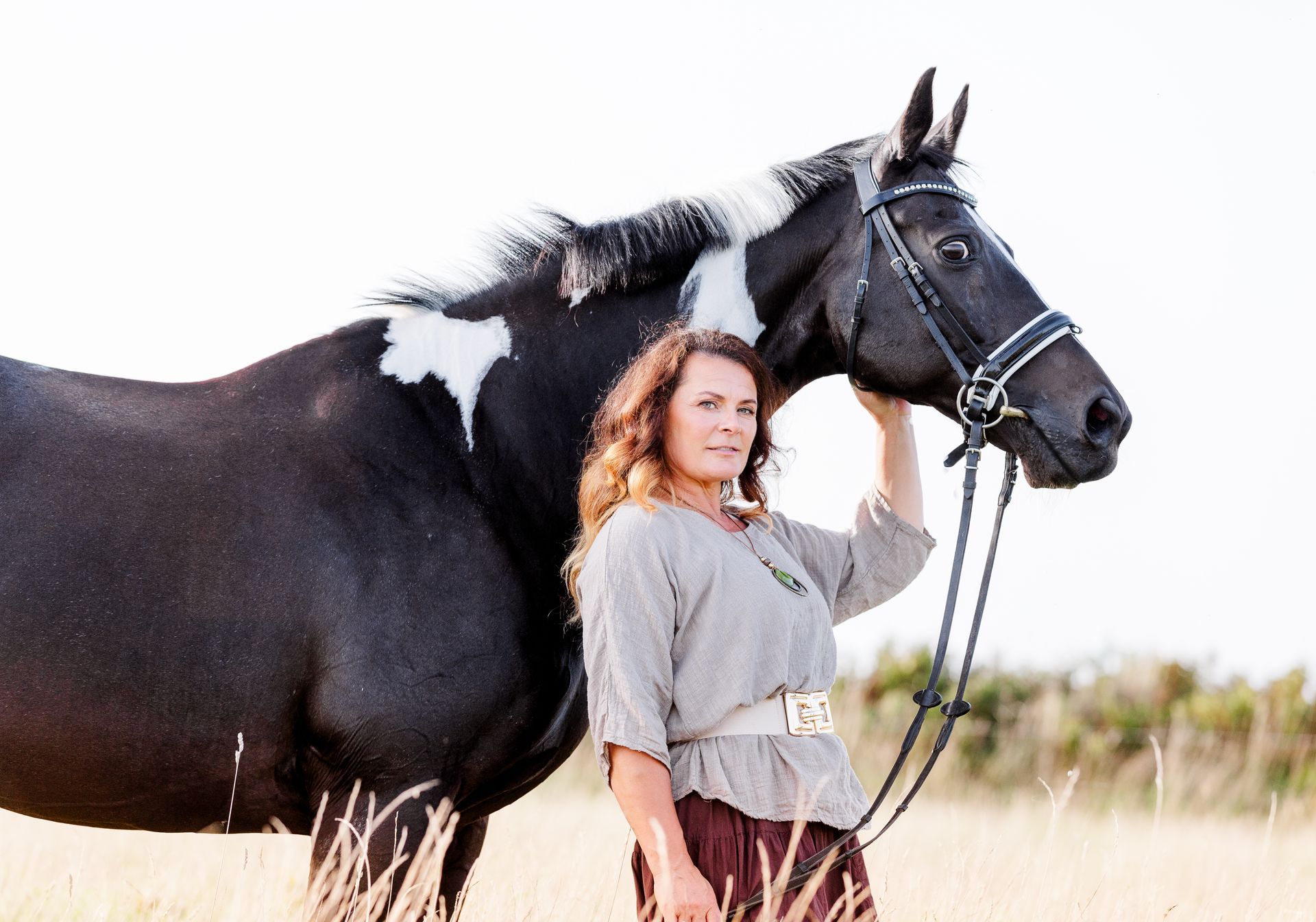 Woman in gray top and brown skirt next to a black and white horse in field.