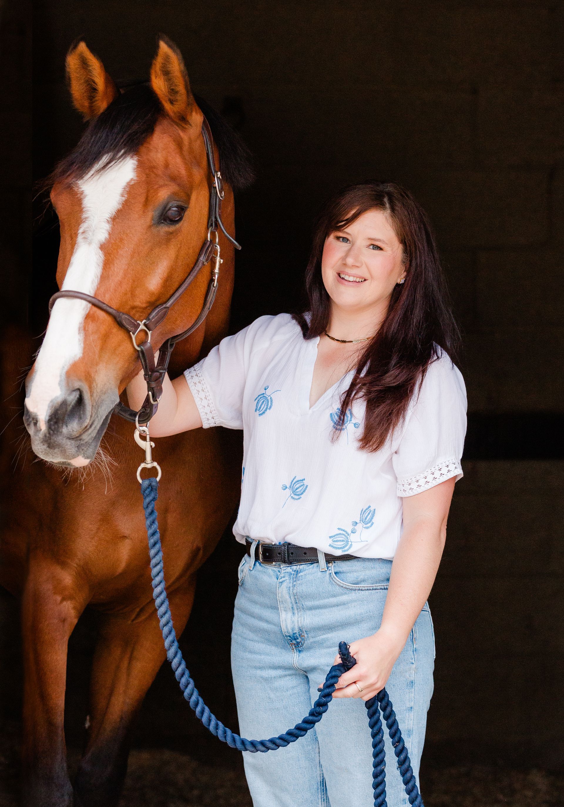 Woman with brown horse, wearing blue jeans and white shirt, in a stable.