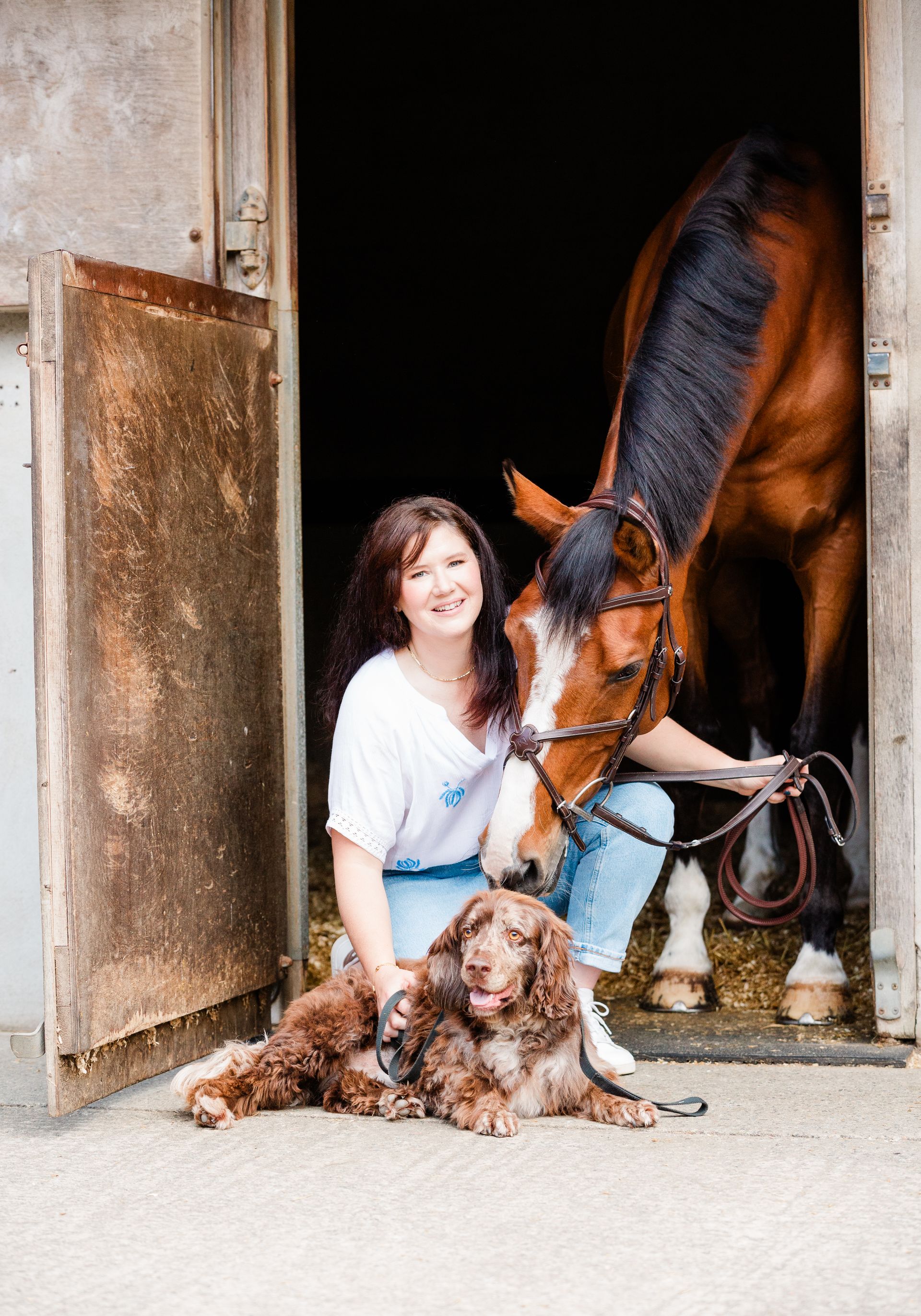 Woman and dog with a brown horse in a stable doorway. The woman is smiling.