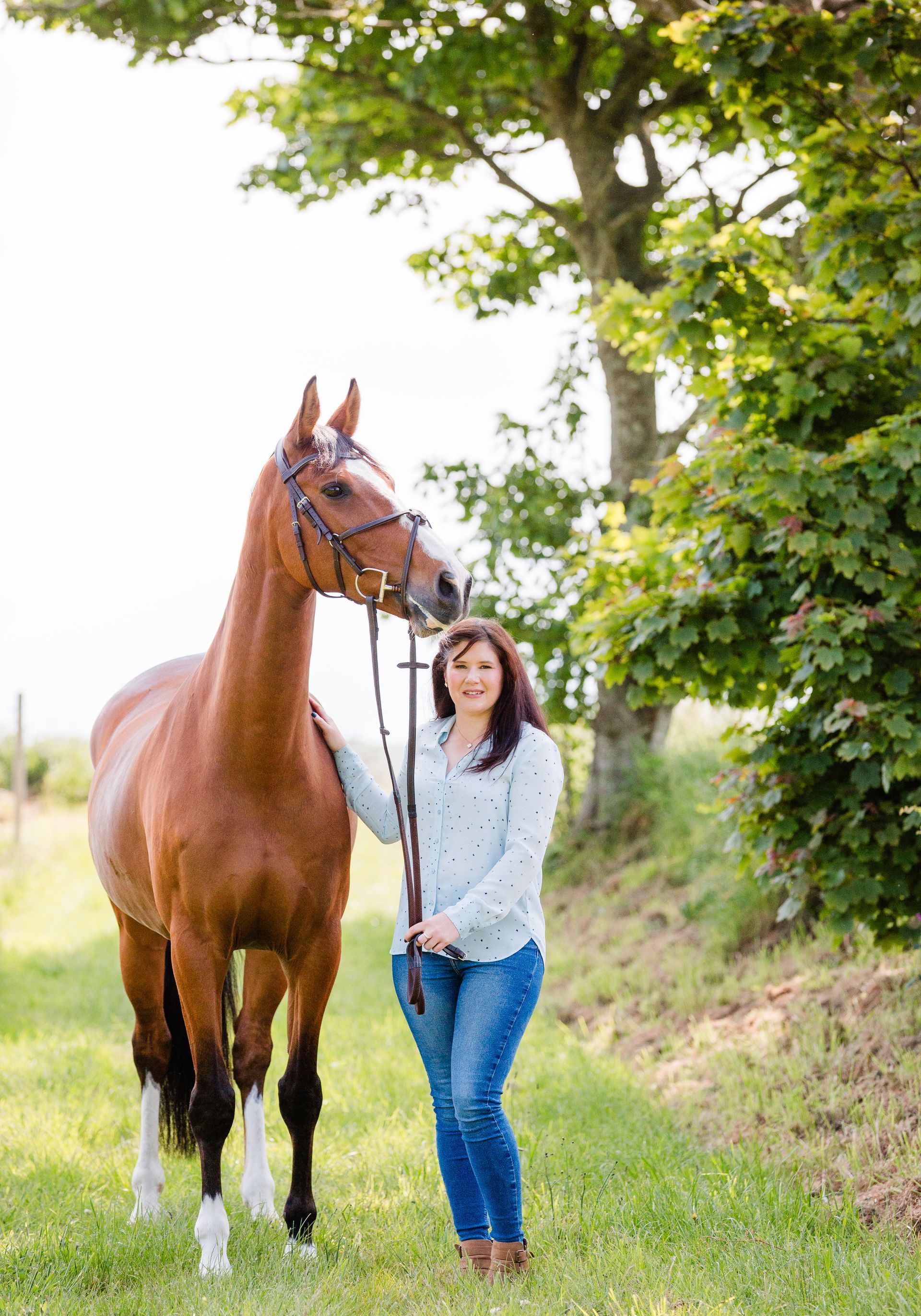 Woman with brown horse outdoors, smiling, holding reins. Green grass, tree in background.