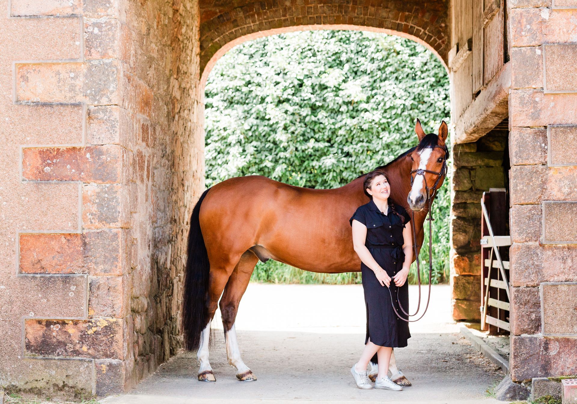 Woman in black dress stands with brown horse under stone archway.