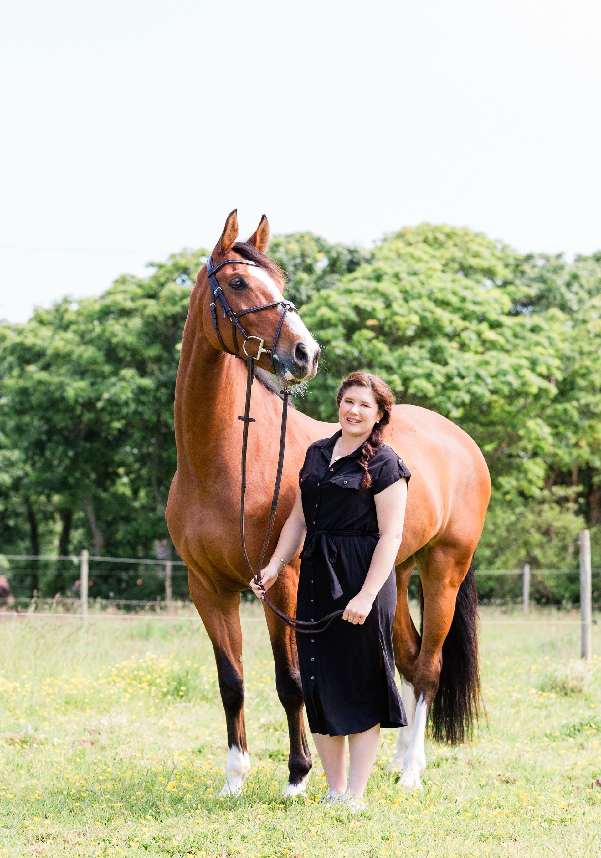 Woman in black dress stands next to a brown horse in a field, holding its reins.