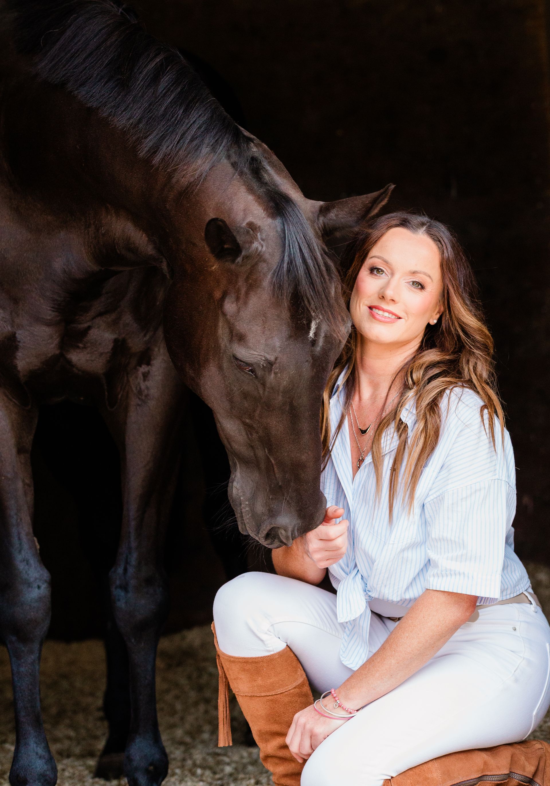 Woman seated with dark horse, petting it. Wearing white pants, tan boots, and a light blue shirt. Barn setting.