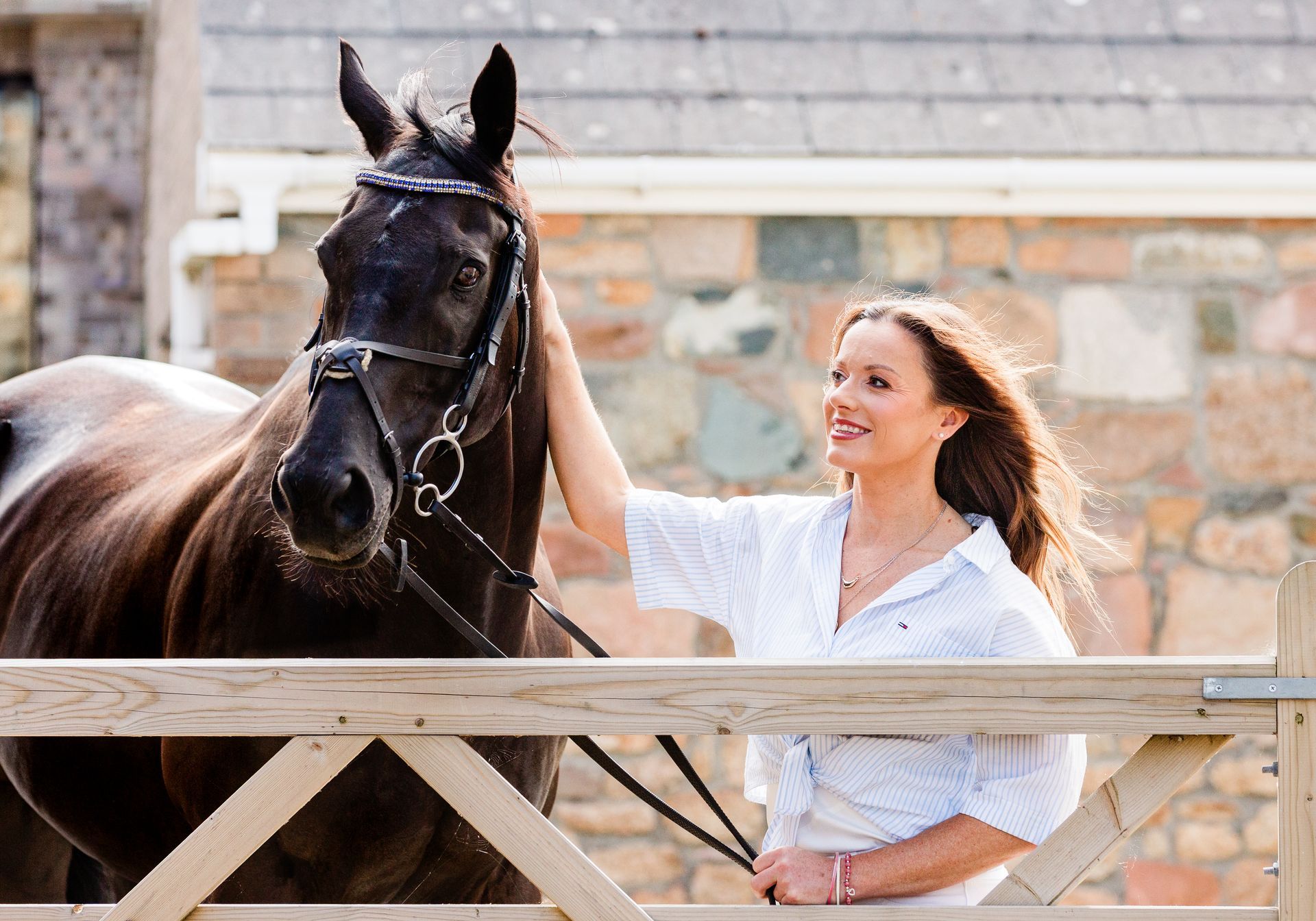 Woman smiles, petting a dark horse. They stand next to a wooden fence. Brick building in the background.