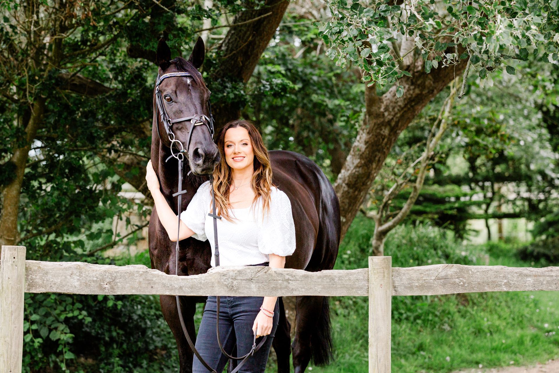 Woman petting a dark horse by a wooden fence, smiling in a natural setting.