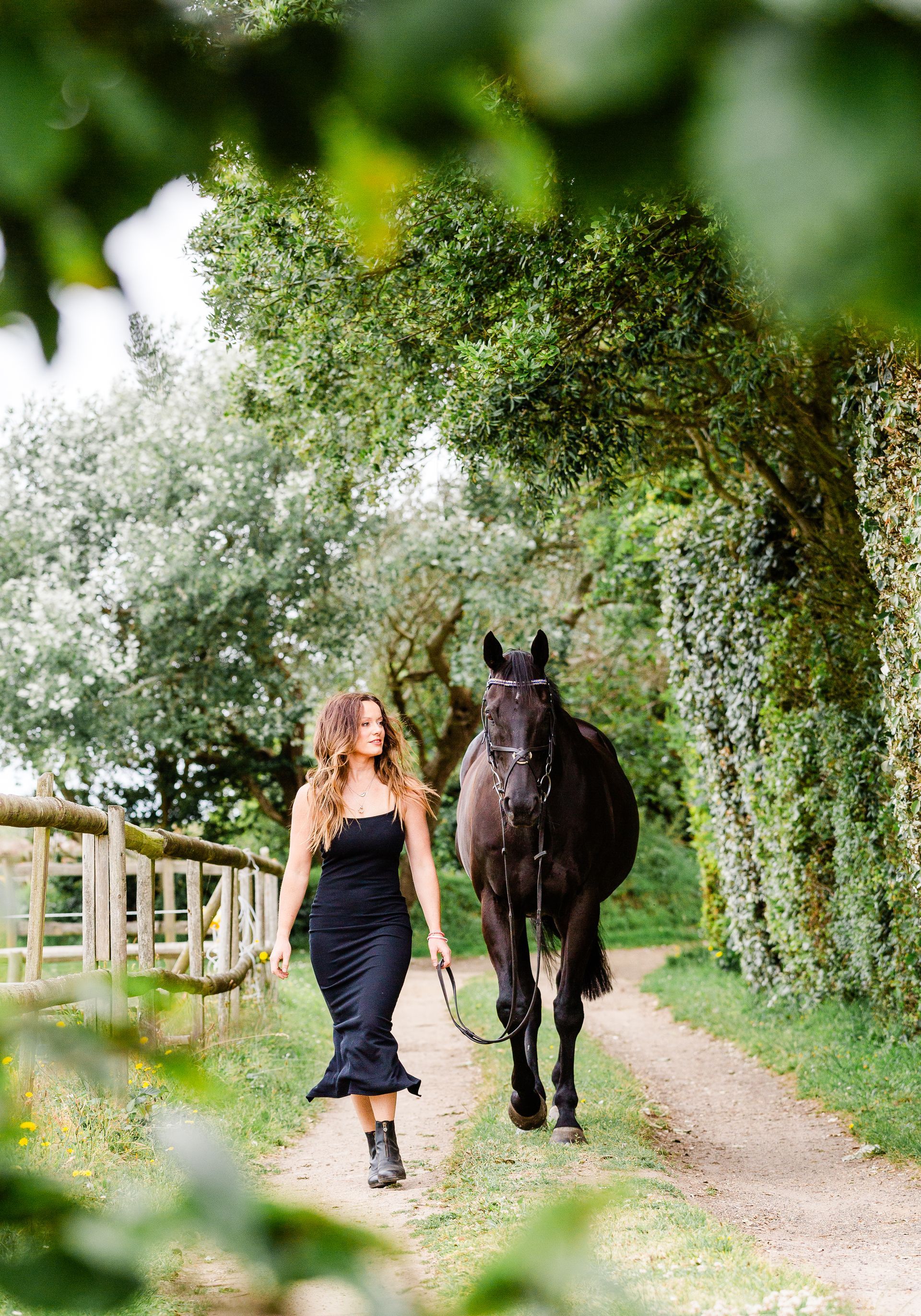 Woman in black dress walks with a black horse on a dirt path lined with trees and a fence.
