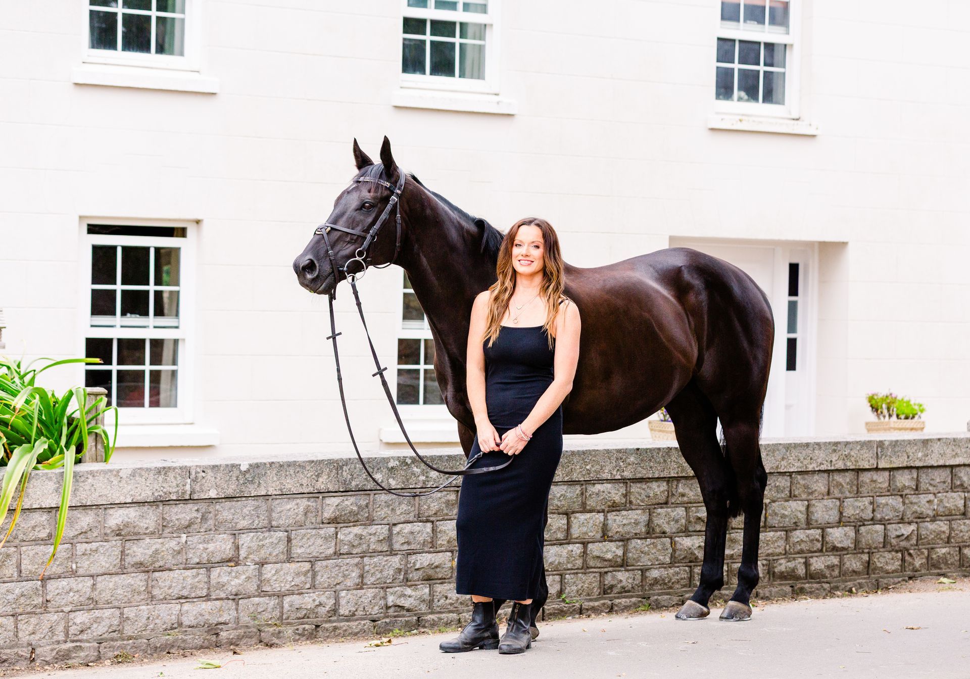 Woman in black dress stands beside a dark horse in front of a white building with stone wall.