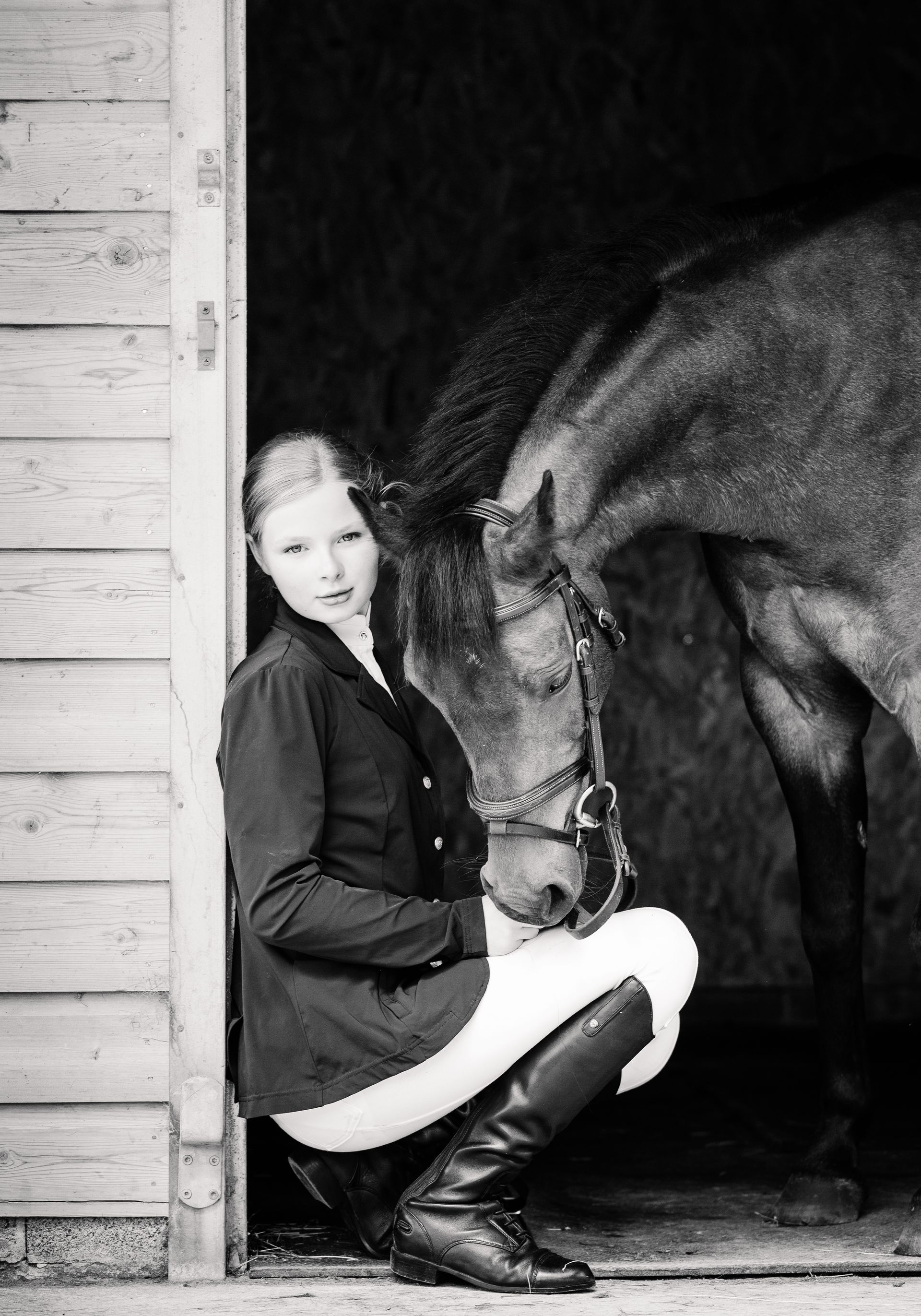 Woman in riding attire crouches next to a horse in a stable doorway.