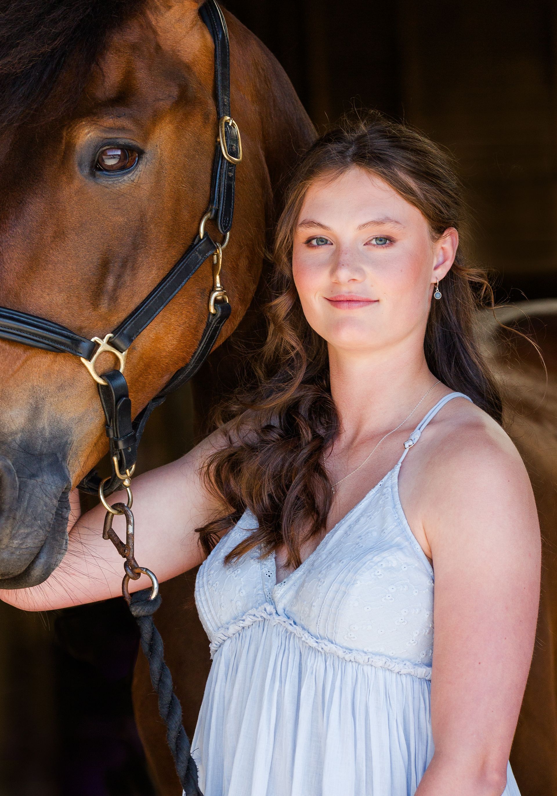 Woman in a light blue dress poses next to a brown horse in a barn.