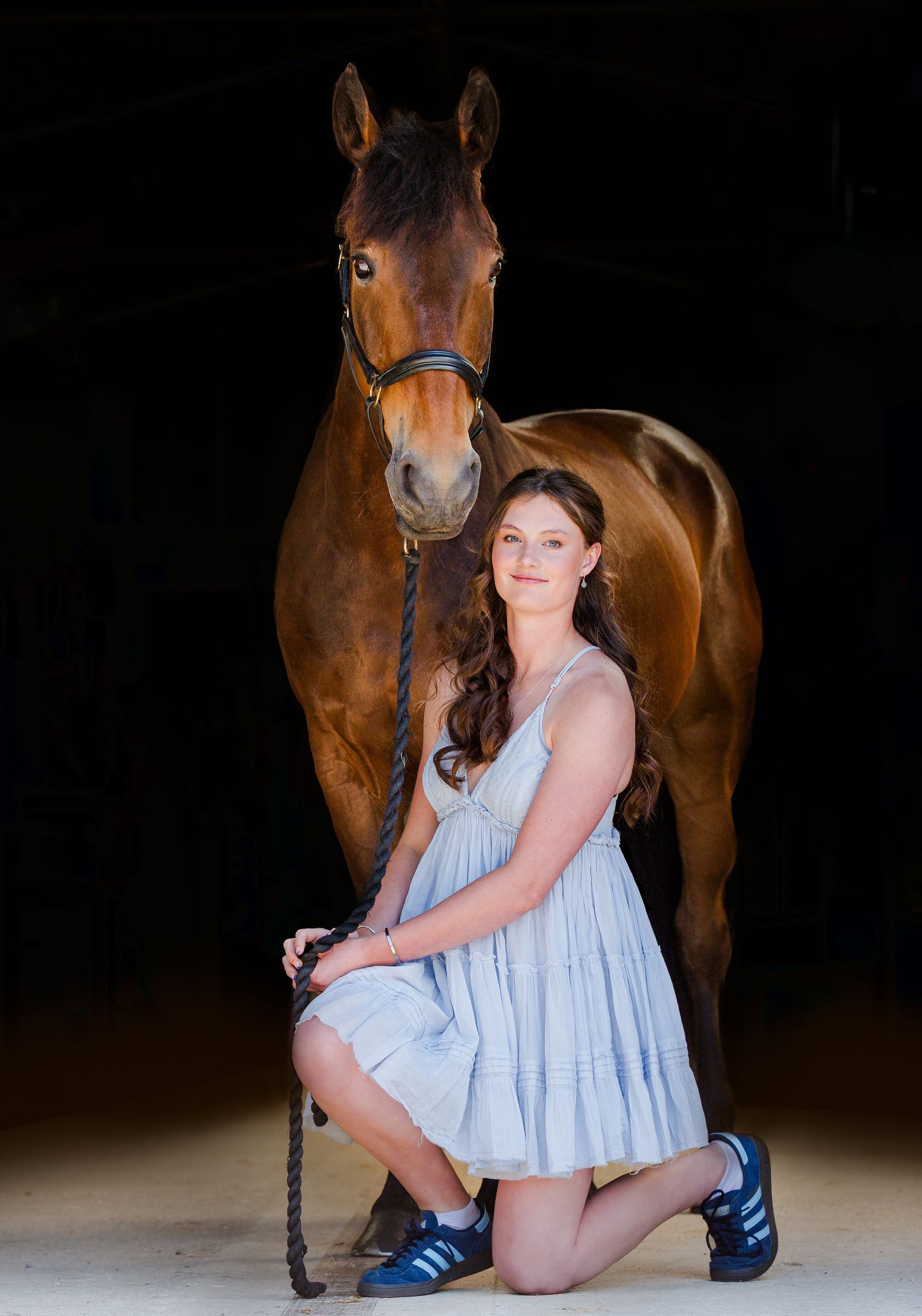 Woman kneeling next to brown horse, posing against a dark backdrop; wearing a blue dress and sneakers.