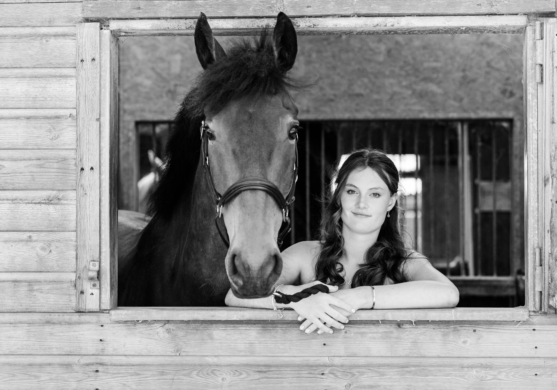 Woman and horse pose together in a stable window; the horse looks forward, the woman smiles with her arms crossed.
