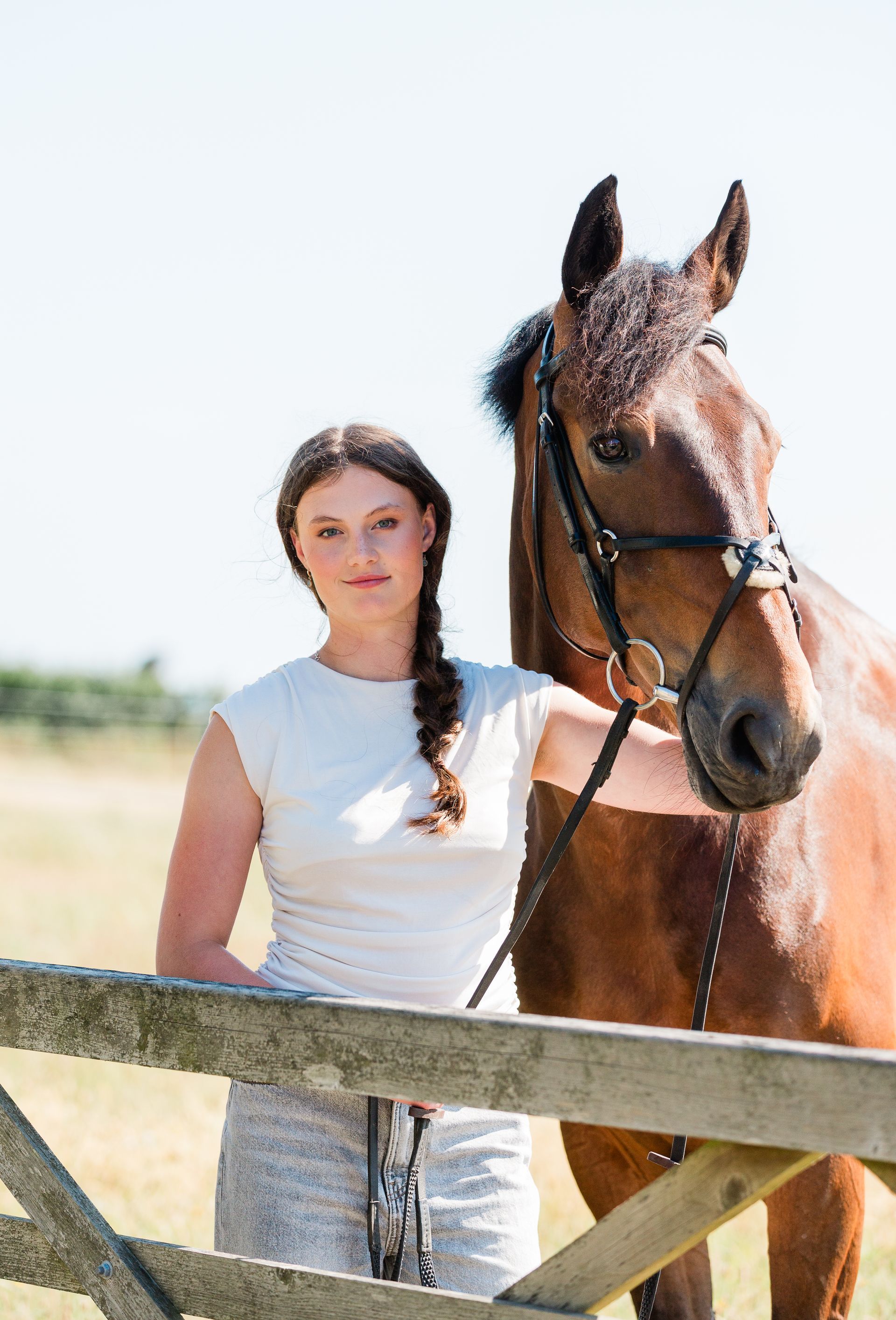 Woman with braided hair leans on a wooden fence, next to a brown horse in a sunny field.