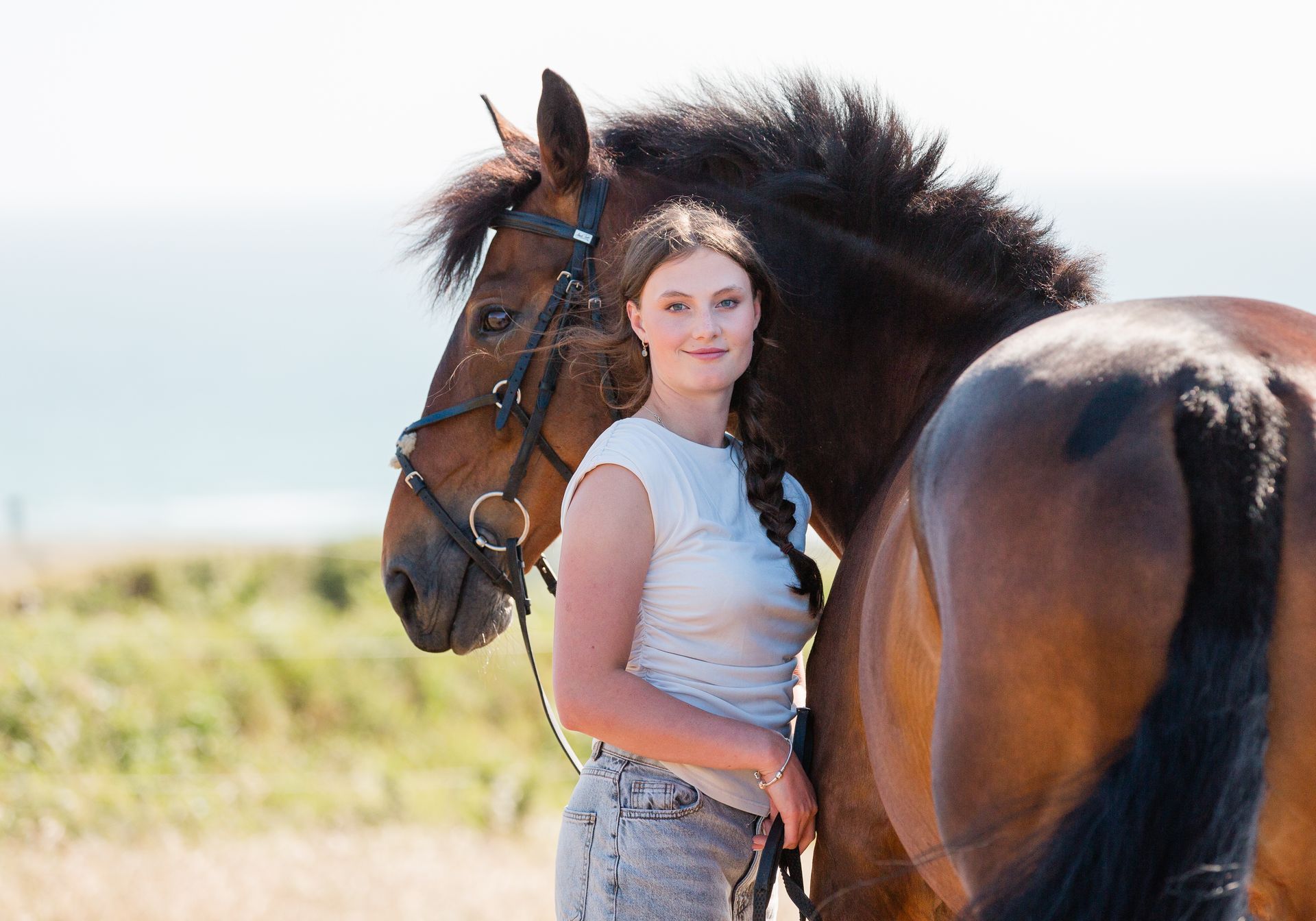 Woman with a brown horse on a sunny day near the coast, woman is wearing a white t-shirt.