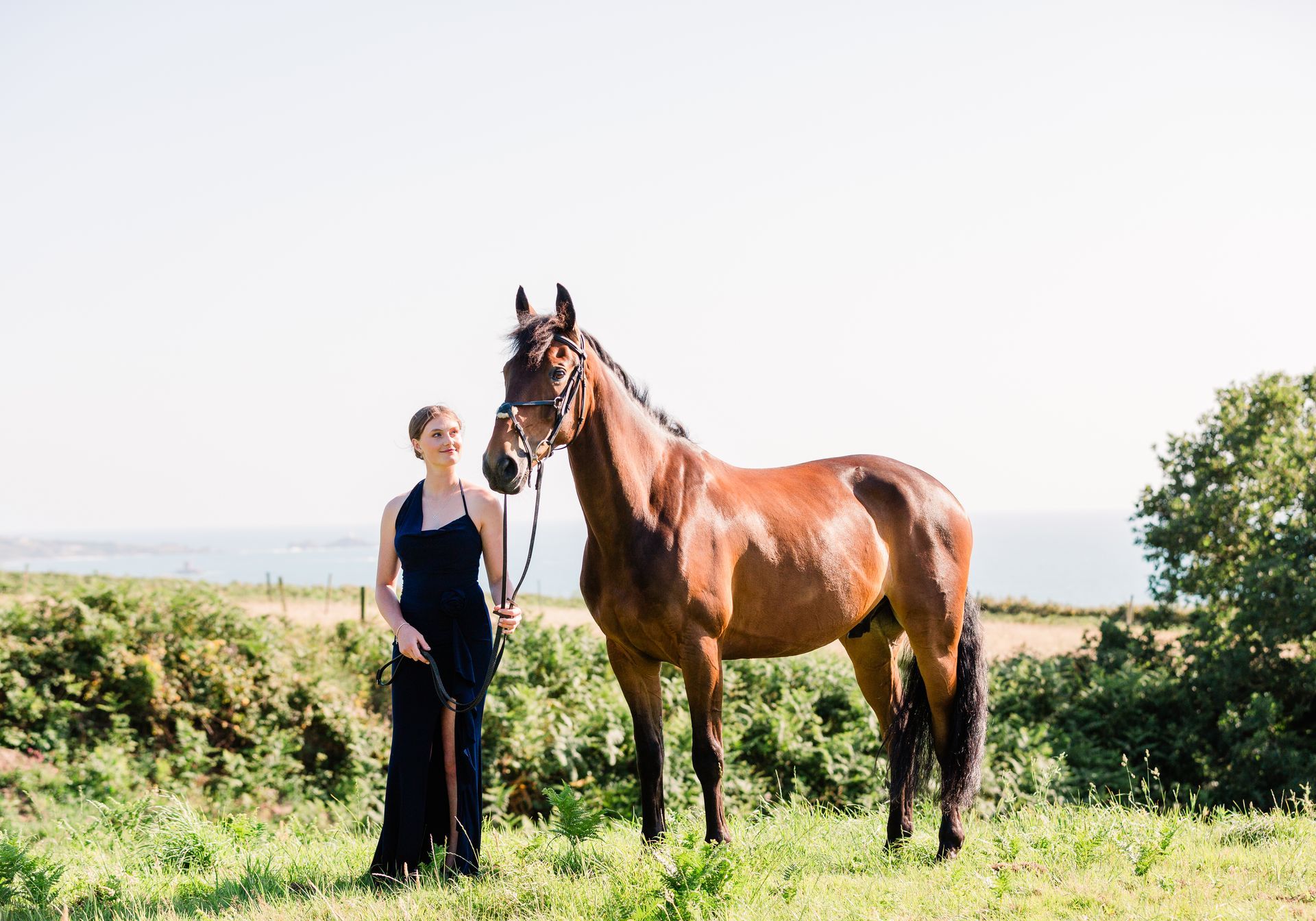 Woman in black dress stands with a brown horse in a field; ocean in the background.