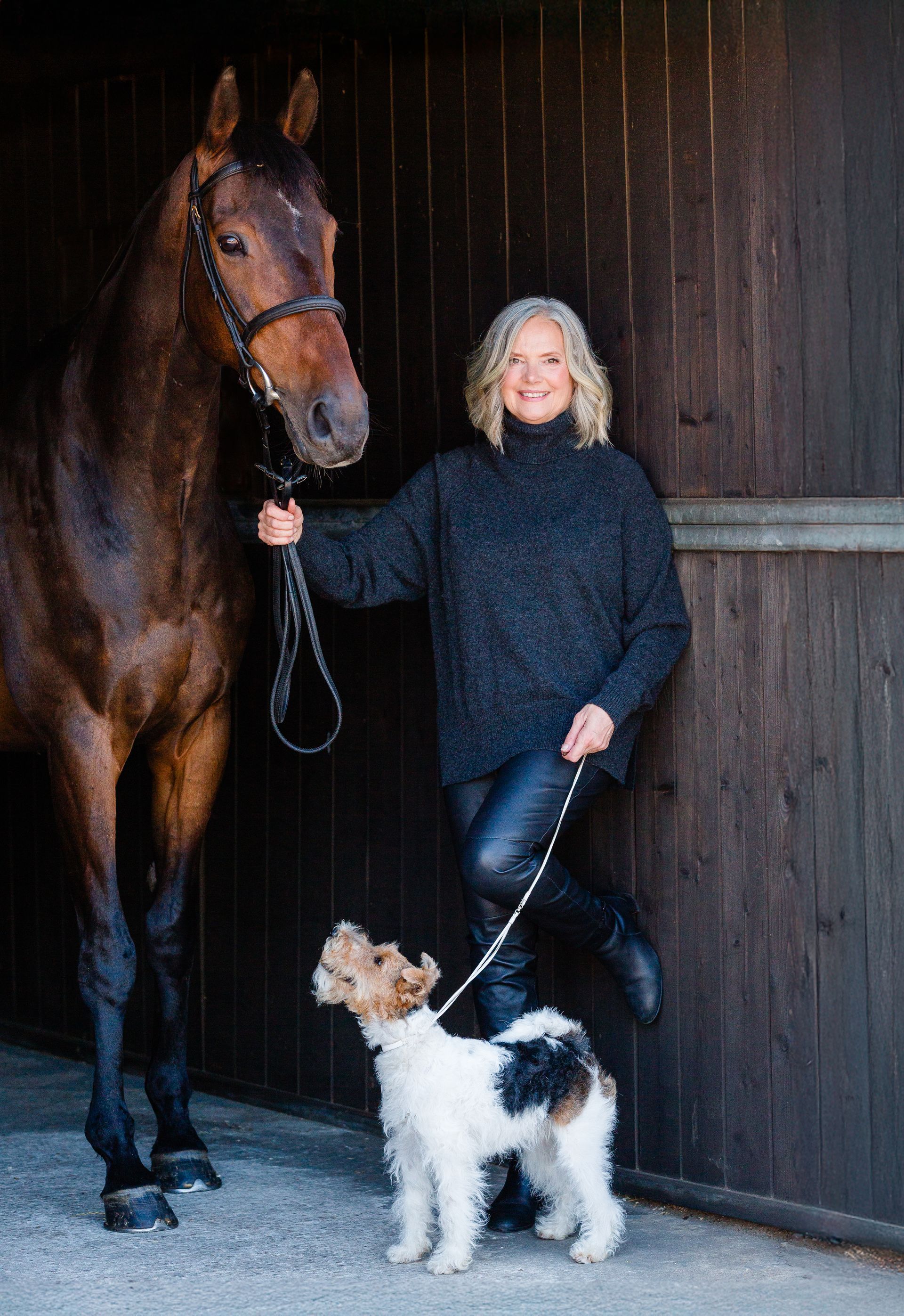 Woman in a dark sweater, holding a horse and dog, inside a stable.
