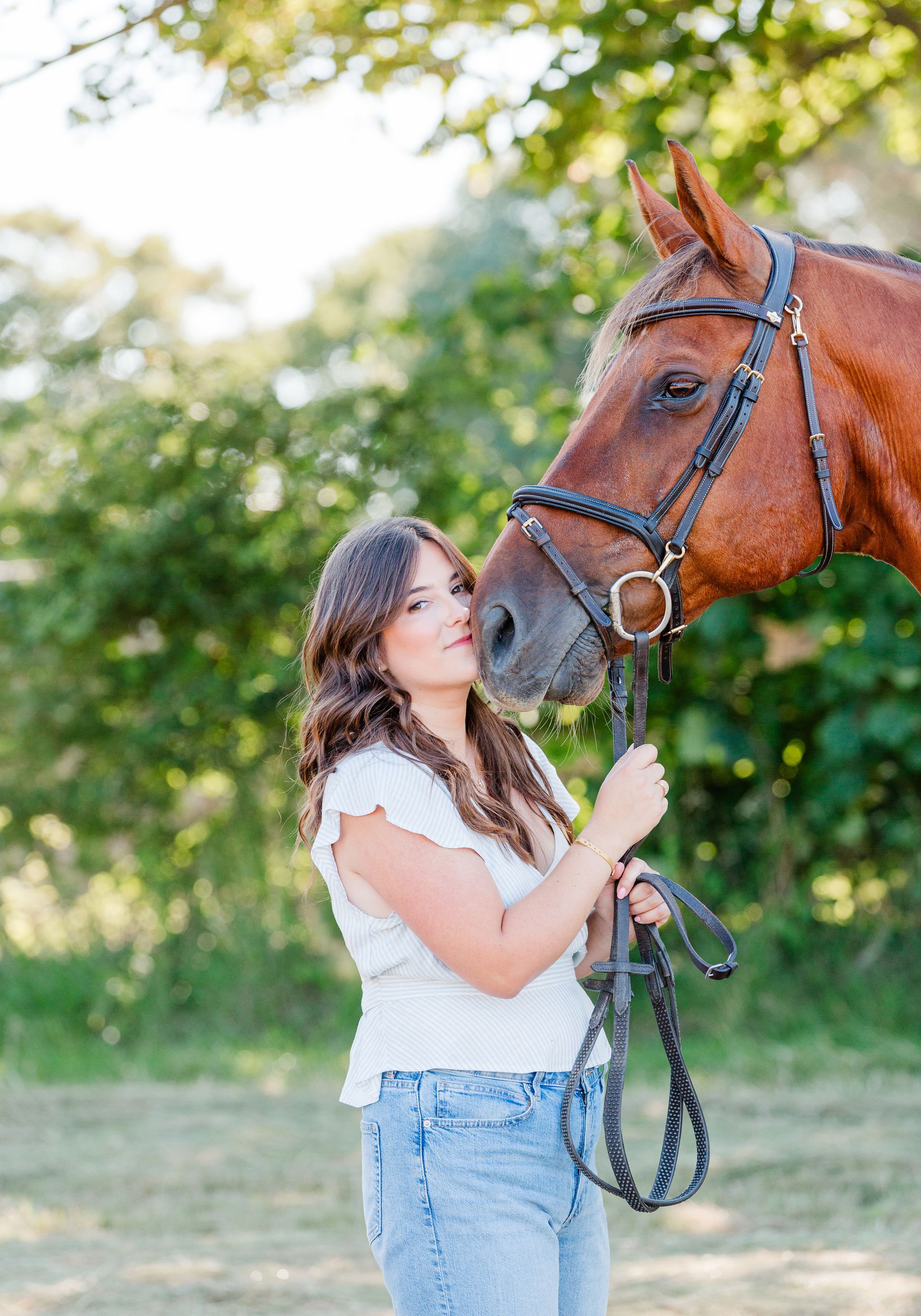Woman with brown horse, outdoors, touching nose, wearing jeans and white top.