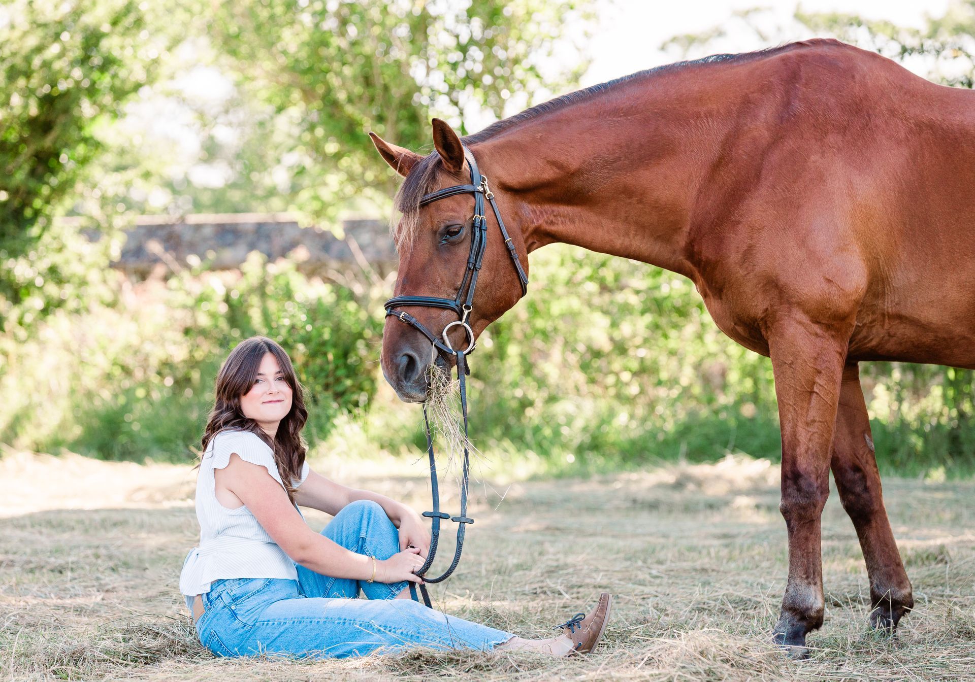 Woman sits on ground next to brown horse, both looking at the camera outdoors.