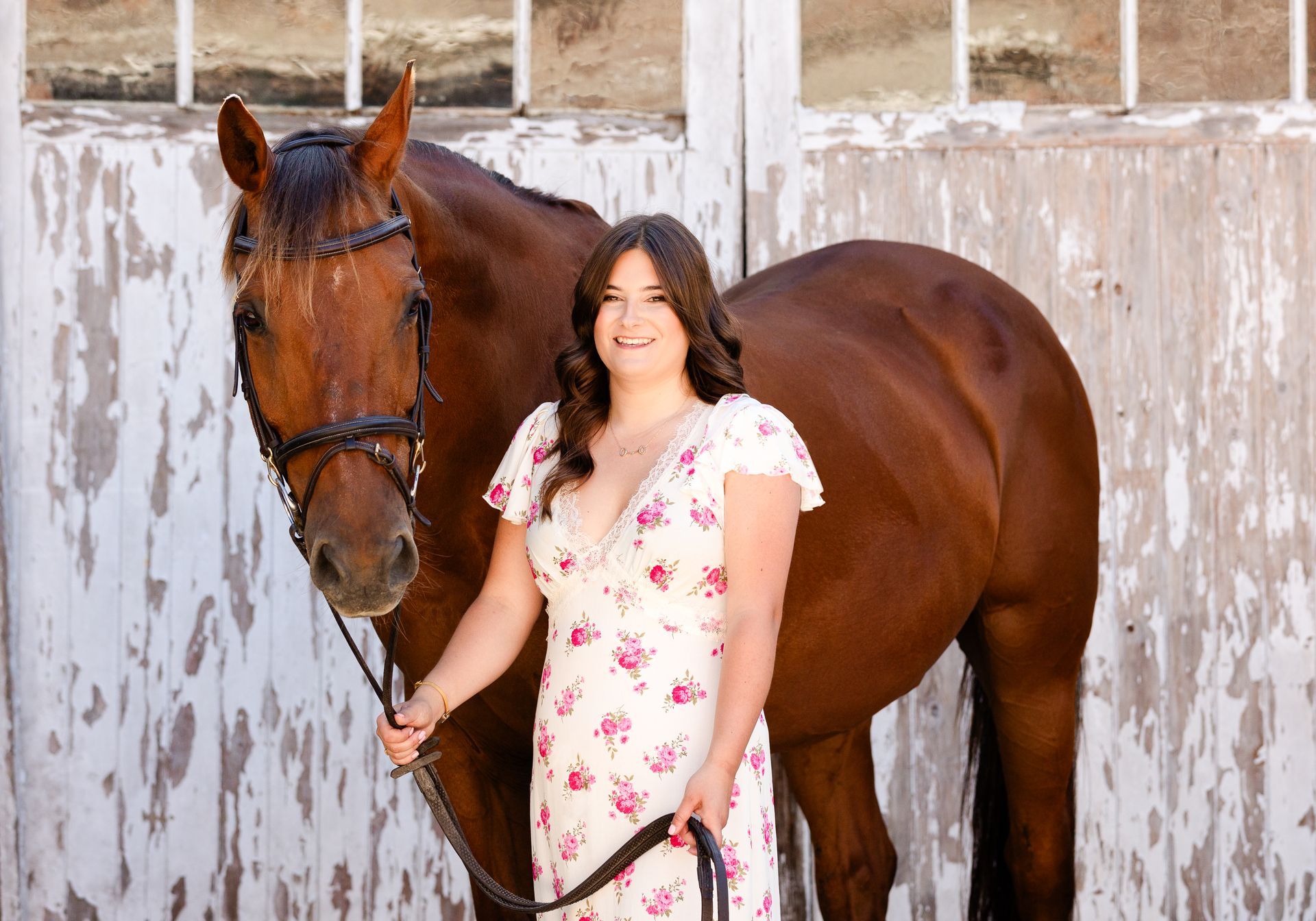 Woman in floral dress with brown horse in front of weathered white barn.