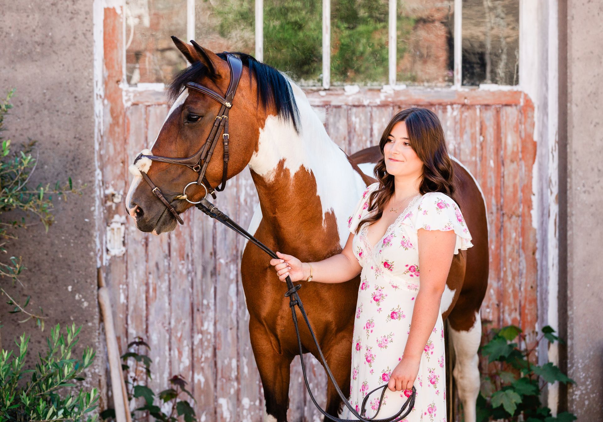 Woman in floral dress holding a brown and white horse by the reins near a weathered wooden structure.