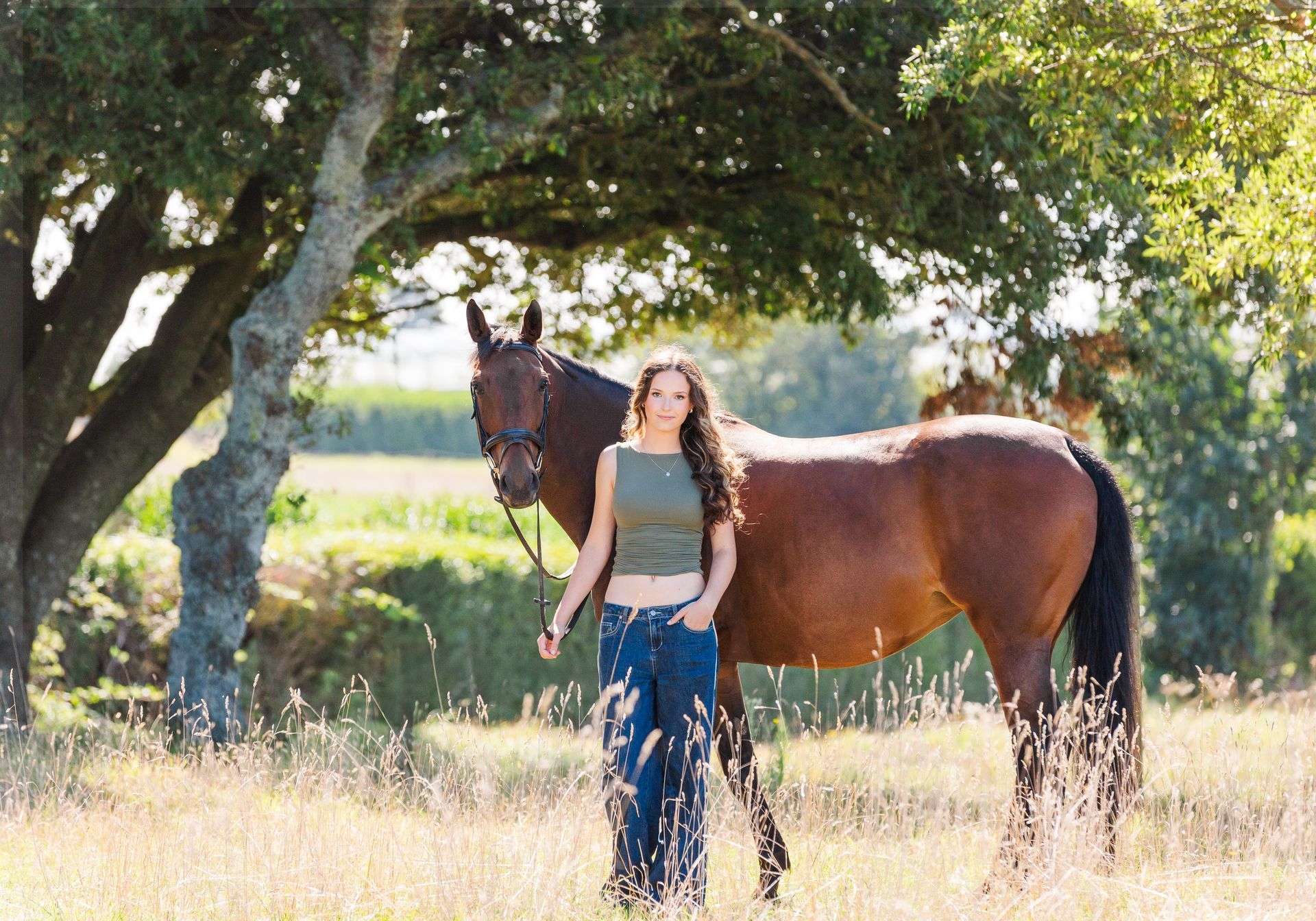 Woman standing with brown horse under a tree in a sunny field.