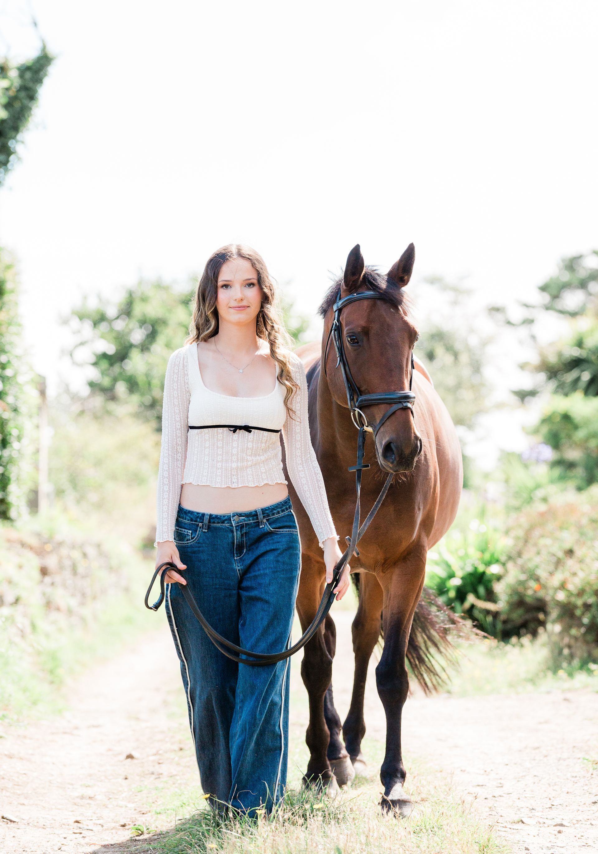 Woman in white top and blue jeans walking a brown horse on a path.
