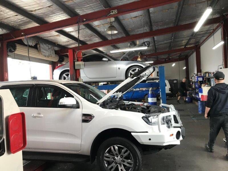 A White SUV With Open Hood and a Silver Car on a Lift in a Garage With People Working — Mike's Automotive in Edgeworth, NSW