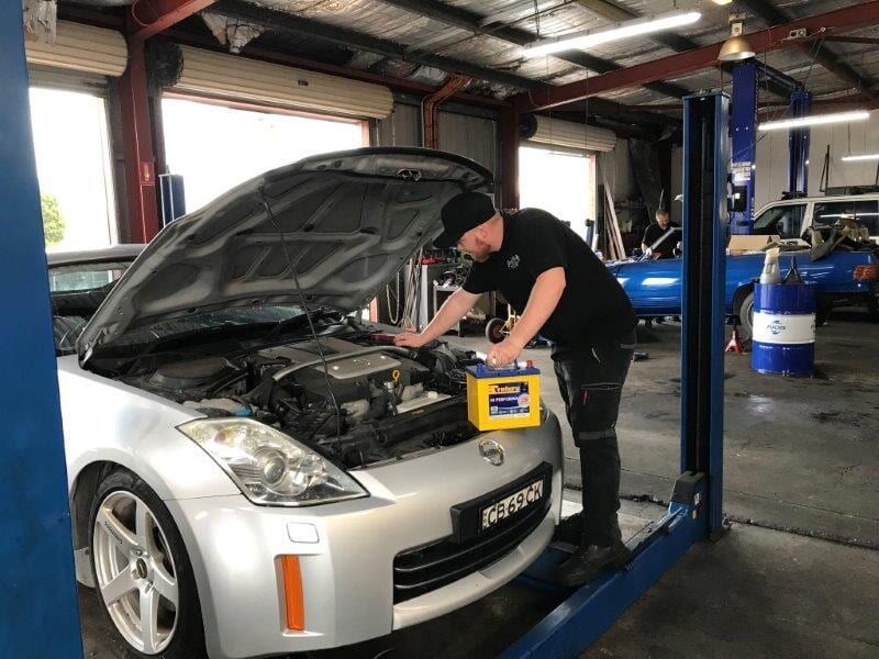 Mechanic Working on a Silver Car in a Garage, Holding a Yellow Battery — Mike's Automotive in Edgeworth, NSW