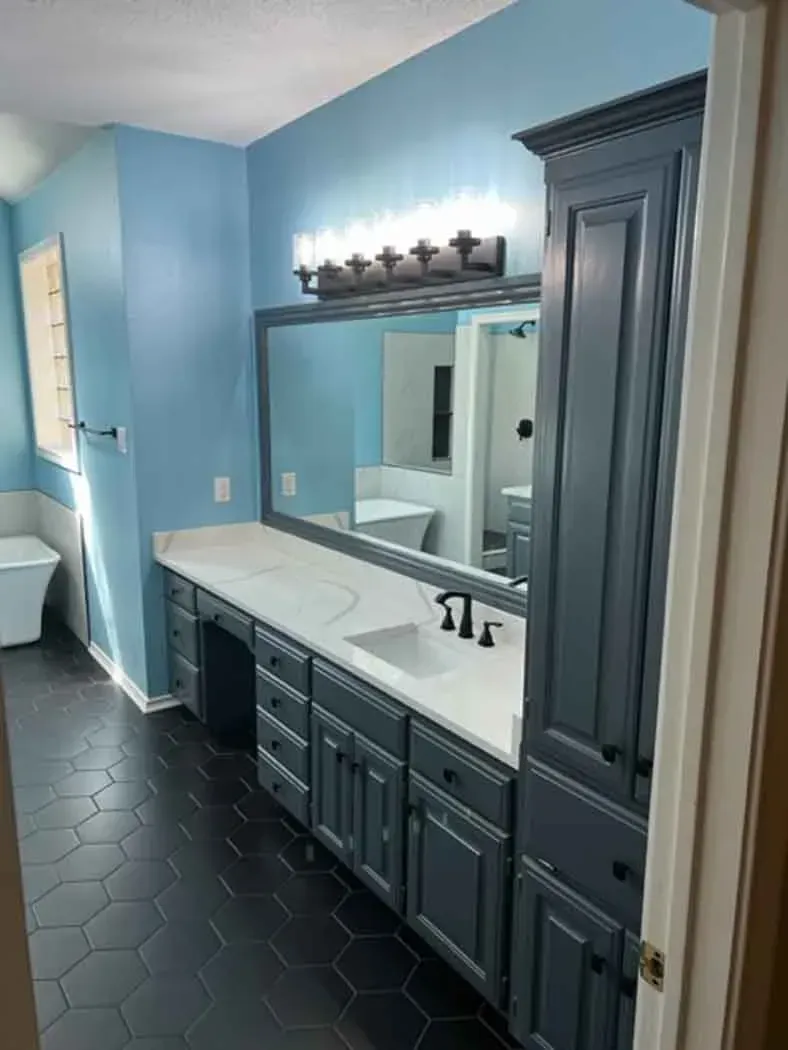 Blue bathroom with dark hexagon tile floor, vanity and cabinet.  Vanity has a large mirror, and the wall is painted light blue.