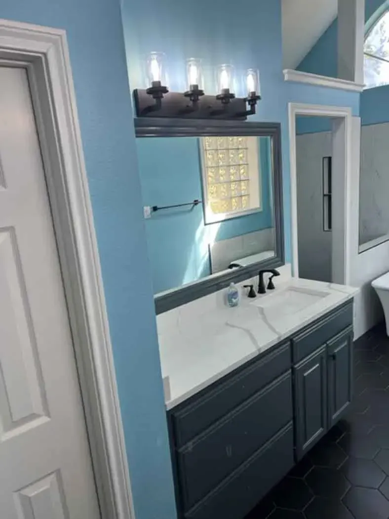 Blue bathroom with a vanity, mirror, and a window. Black flooring, white door, and gray cabinets.