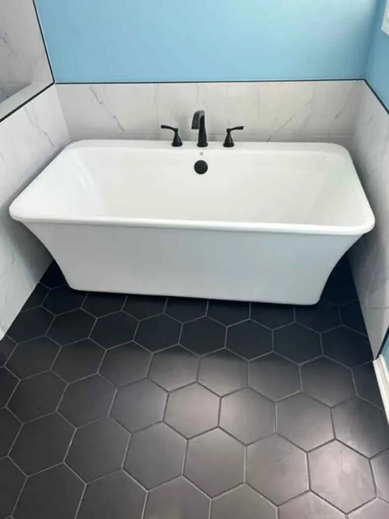 White freestanding bathtub with black fixtures, set on black hexagonal tile, against blue and white walls.