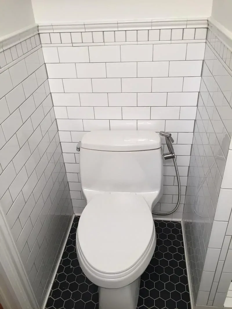 White toilet in a small bathroom with white tiled walls and black hexagon floor tiles.