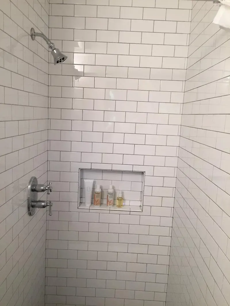 White-tiled shower with a built-in shelf holding toiletries; chrome showerhead and faucets.