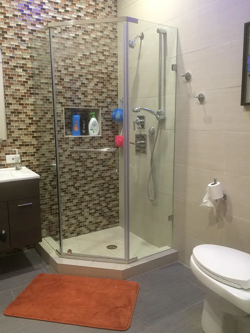 Modern bathroom with glass shower, toilet, vanity, and orange bath mat.