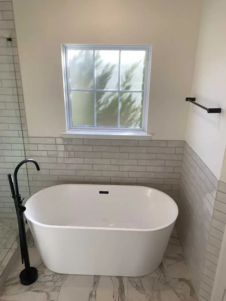 Freestanding white bathtub in a bathroom, under a frosted window and tile backsplash.