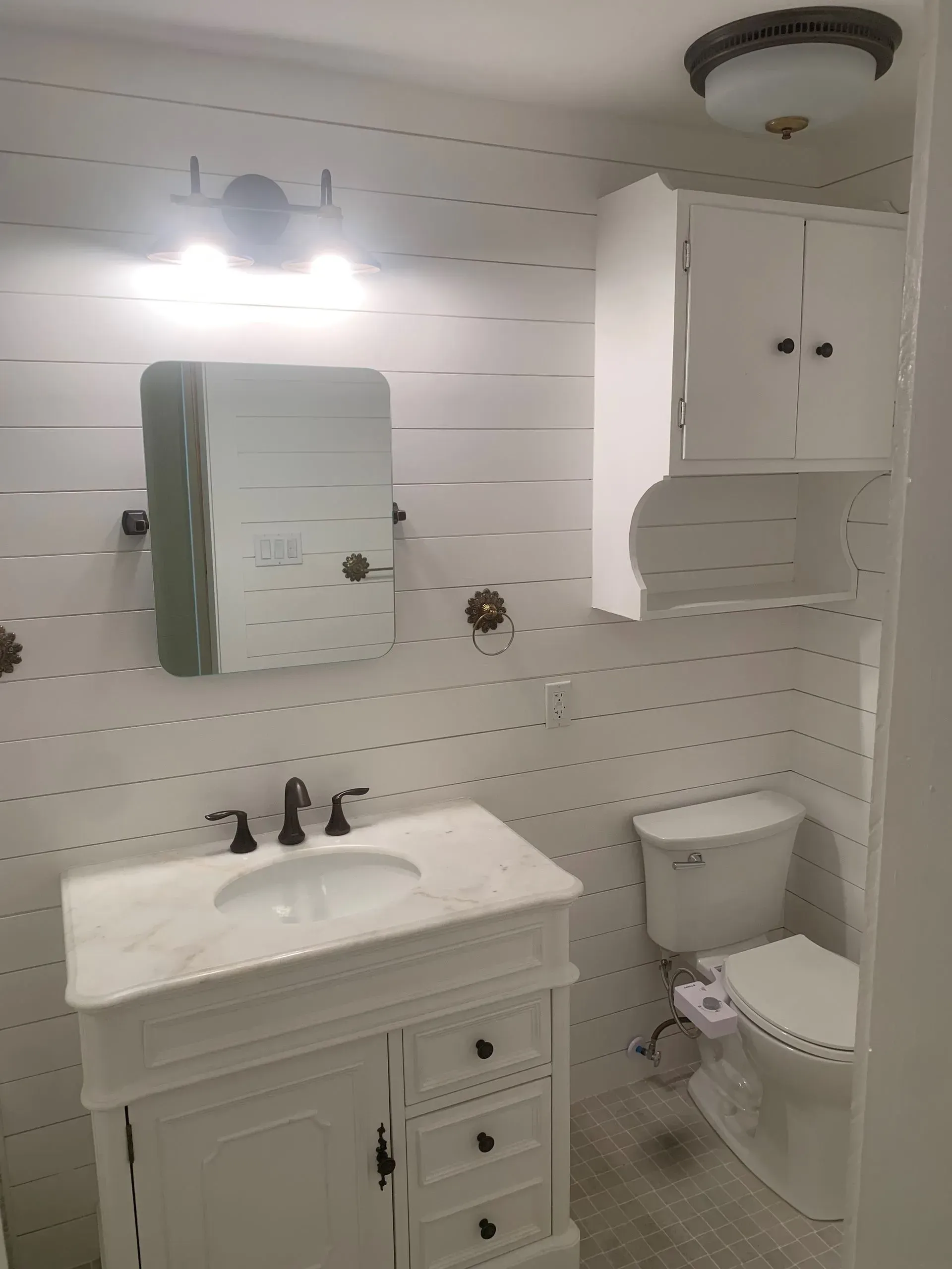 White bathroom with marble vanity, mirror, toilet, and cabinet on shiplap walls.