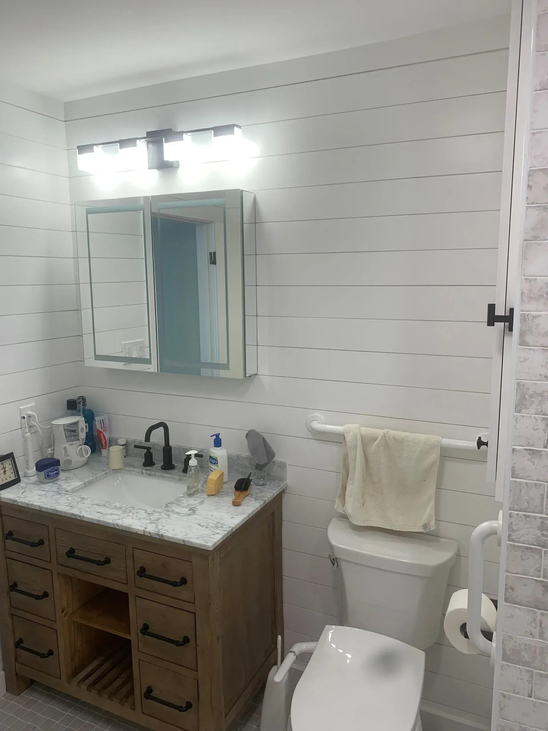Bathroom with white plank-style walls, wooden vanity with granite countertop, toilet, and lit mirror.