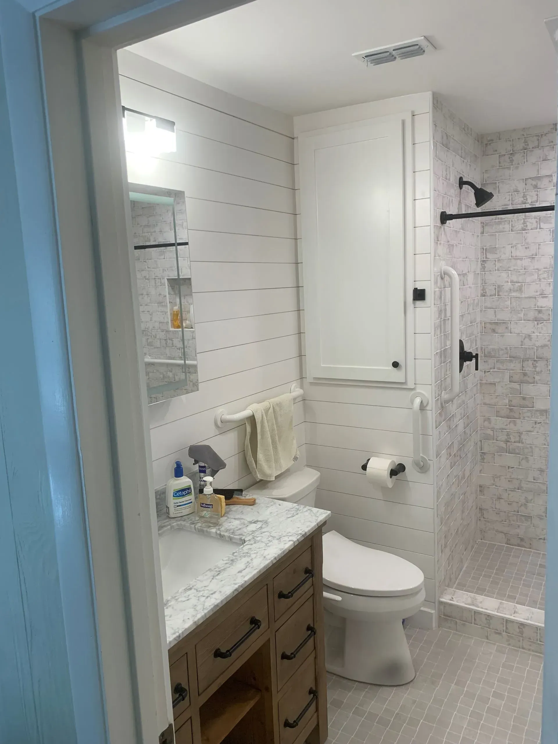 Bathroom with white shiplap walls, wood vanity, shower with mosaic tile, and tall white storage cabinet.