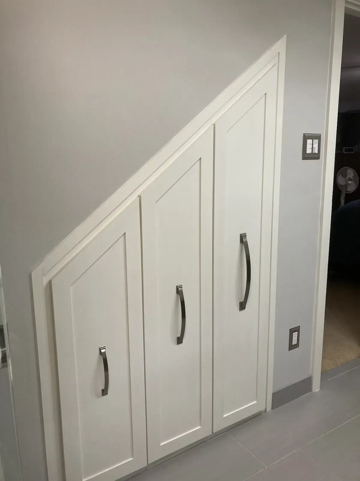 White built-in storage cabinets under a staircase with dark handles. Gray walls and flooring.