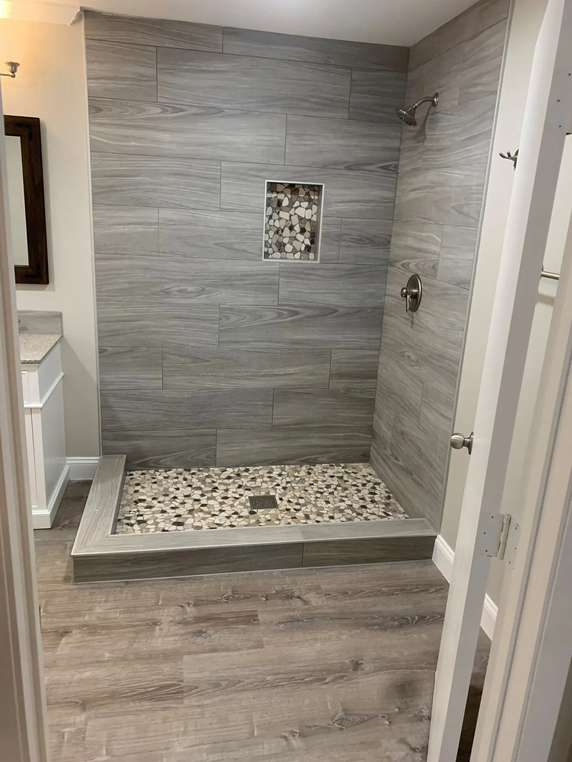 Modern bathroom with gray tiled shower and pebble floor, wooden floor, and open door.