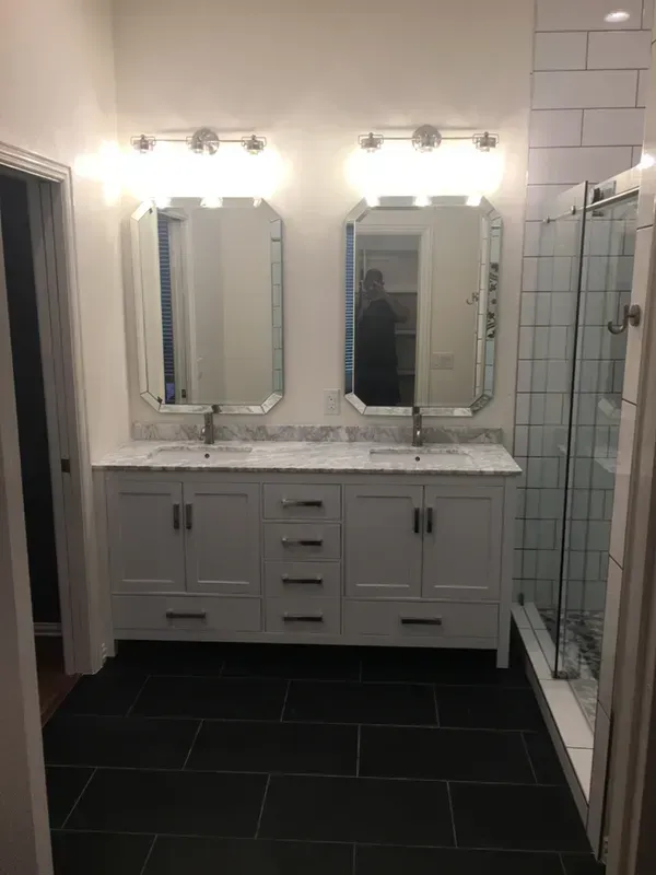 White double sink vanity with two mirrors and lights, black tile floor, and a glass shower.