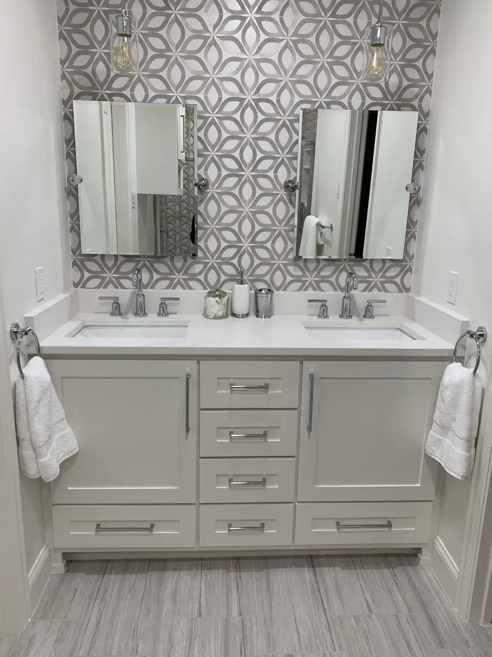 White double vanity with grey floral tile backsplash and two mirrors.
