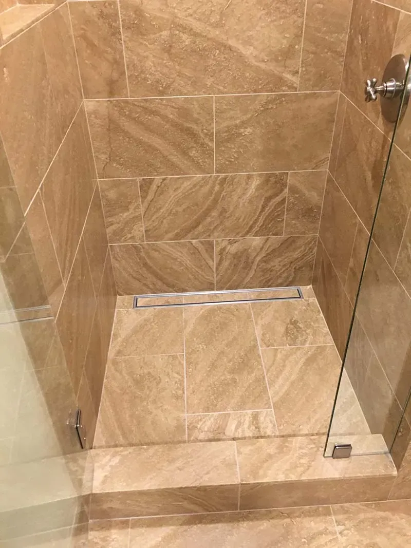Shower stall with beige tile, glass door, and linear drain.