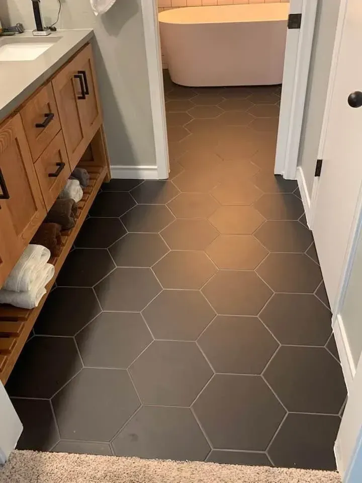 Black hexagon tile floor in a bathroom, leading to a bathtub. Wooden vanity and white towels are visible.