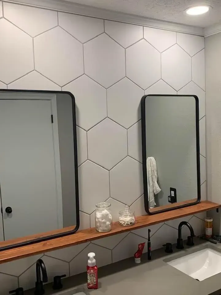 Bathroom with hexagon tile wall, two black-framed mirrors, wooden shelf with toiletries, and dark fixtures.