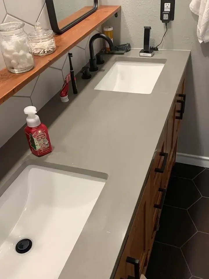 Bathroom with double sinks, gray countertop, wooden vanity, black fixtures, and honeycomb tile floor.