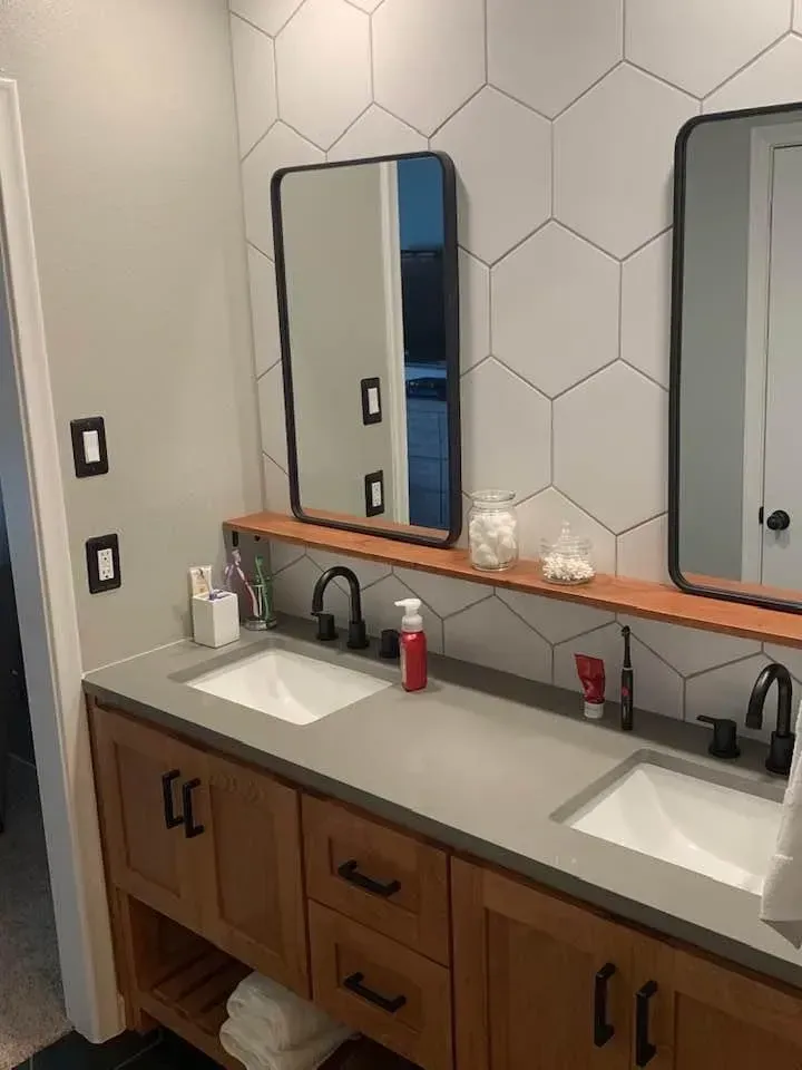 Bathroom with double sinks, wooden vanity, gray countertop, hexagon tile wall, black framed mirrors, and black faucets.