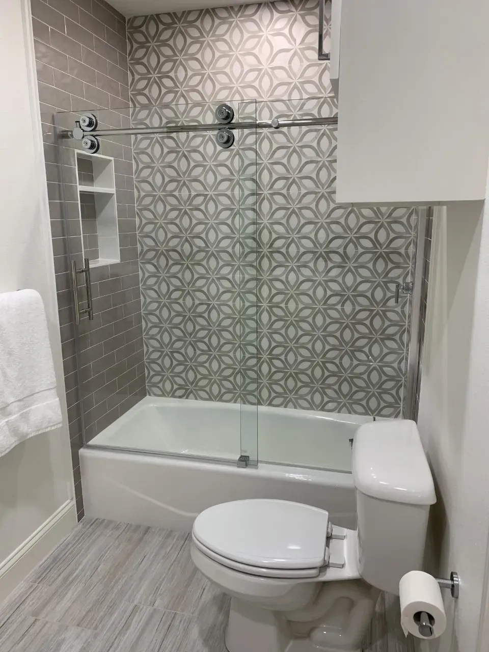 Bathroom with white tub and toilet, patterned gray wall tiles, and glass shower door.
